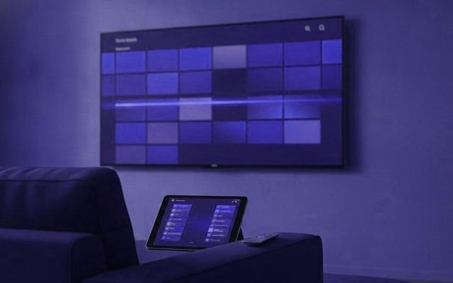 Television showing a programme guide grid above a sofa, with a tablet listing channels propped on the armrest