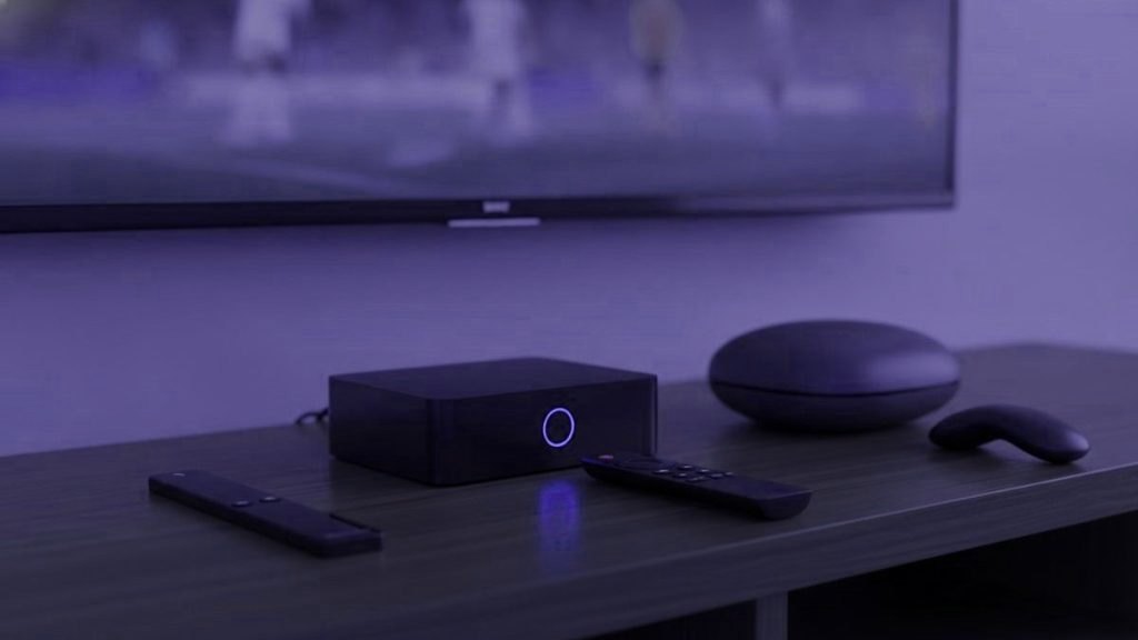 Black set-top box with a glowing ring, a round streaming puck and three remotes on a TV cabinet beneath a television