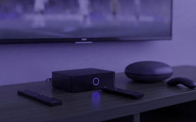 Black set-top box with a glowing ring, a round streaming puck and three remotes on a TV cabinet beneath a television