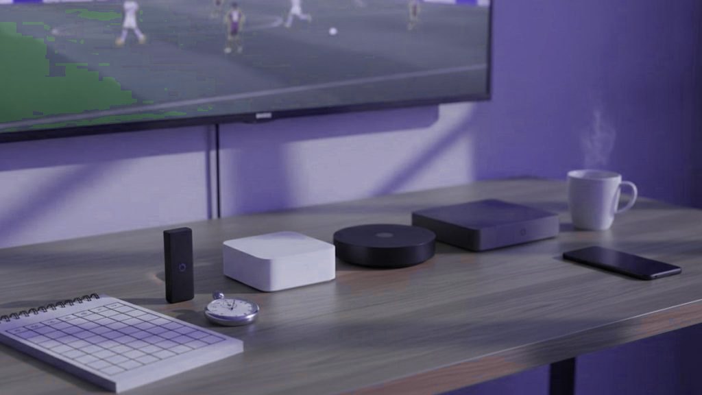 Several streaming devices, a squared notepad, a stopwatch and a mug on a desk beneath a television showing a football match