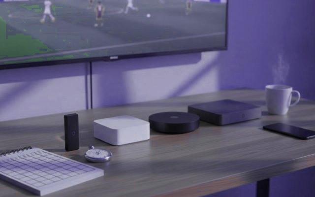 Several streaming devices, a squared notepad, a stopwatch and a mug on a desk beneath a television showing a football match