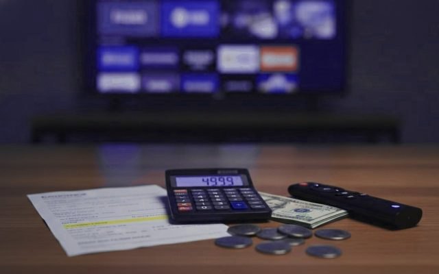 Calculator, banknotes, coins, a TV remote and a printed bill on a wooden table in front of a television menu